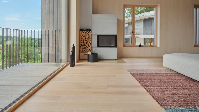 mafi Naturholzboden Esche in einem modernen Wohnzimmer. Links mit Balkon ins Grüne, mittig ein Ofen und rechts einem Sofa